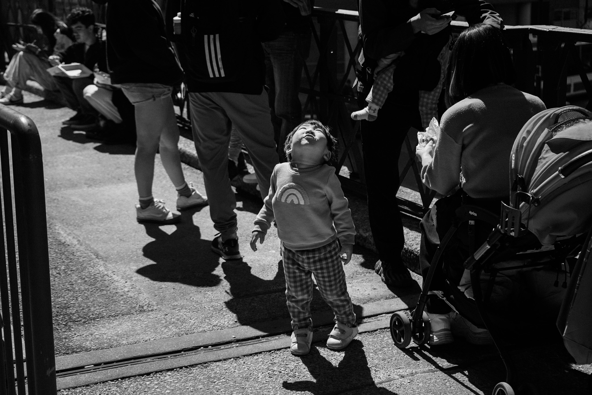 Toddler in a rainbow sweater looking straight up, surrounded by adult legs