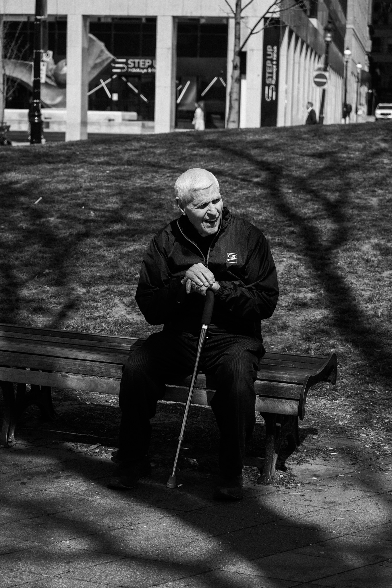 Elderly man on a bench with a cane, city buildings behind him