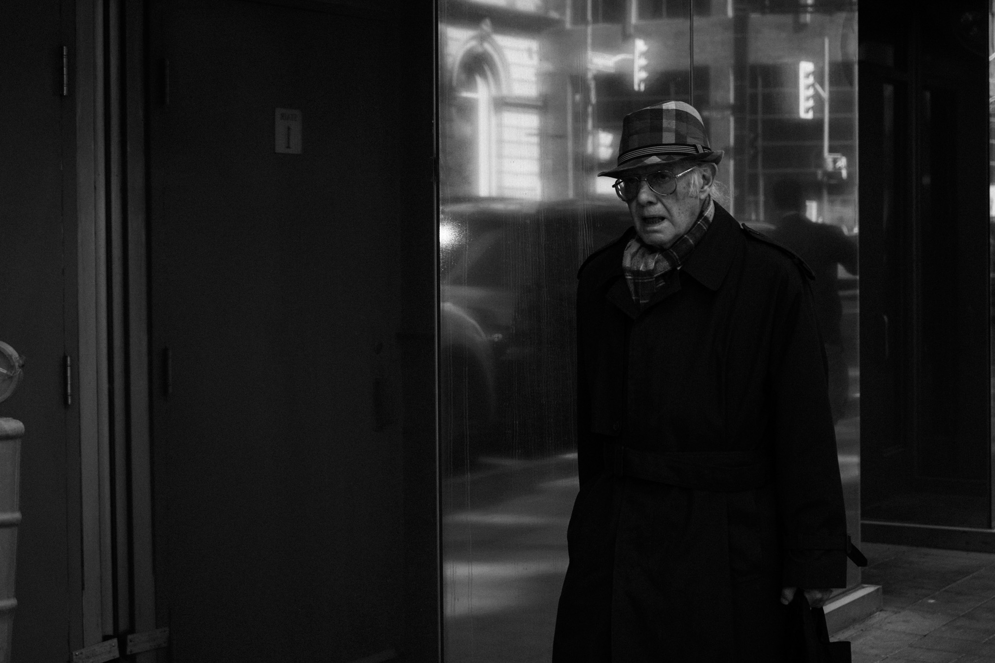 Elderly man in a hat and trenchcoat standing by a glass storefront at dusk