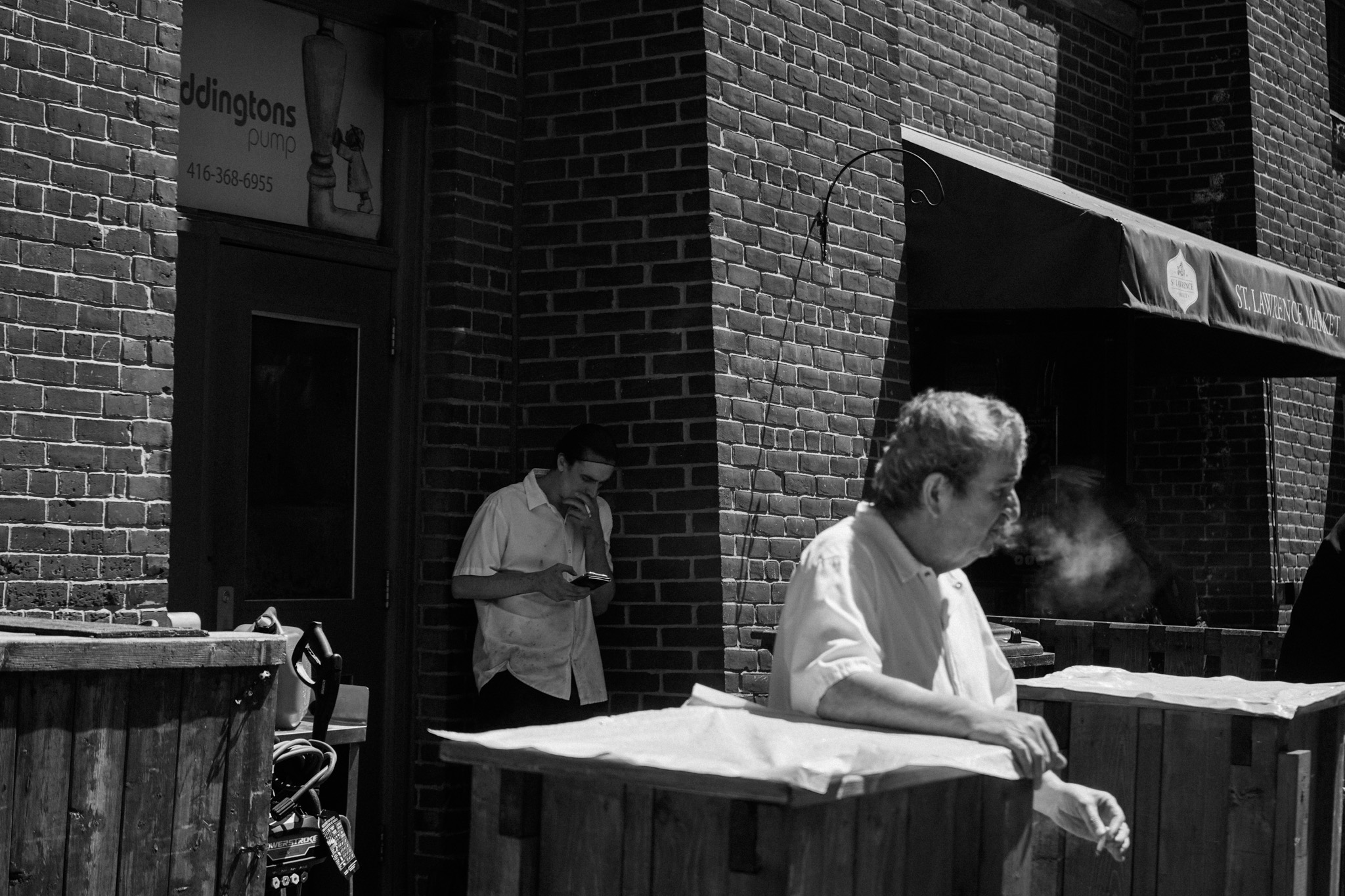 Man smoking outside St. Lawrence Market, another man working in the foreground