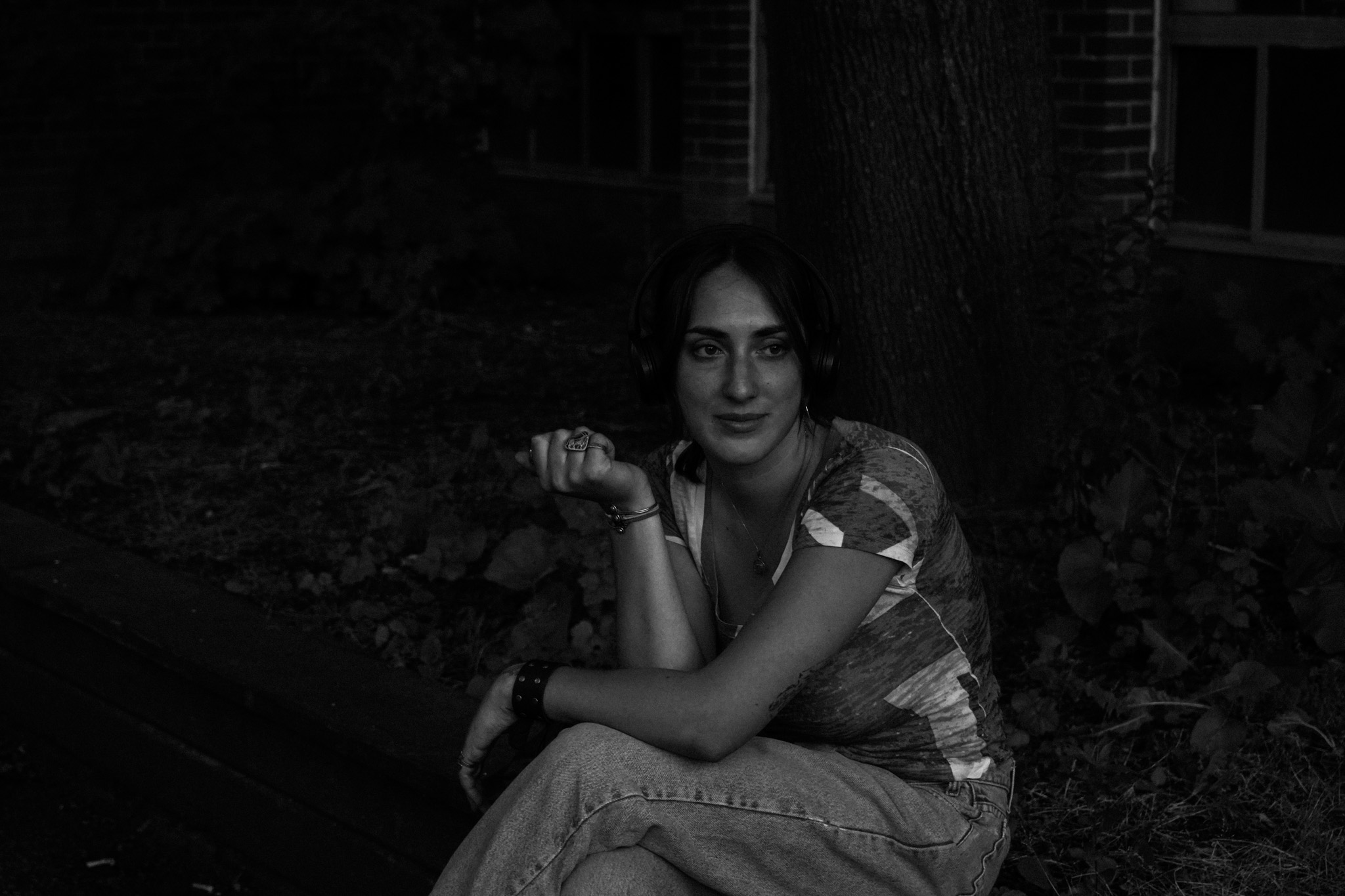 Young woman sitting under a tree at night wearing headphones, looking at the camera