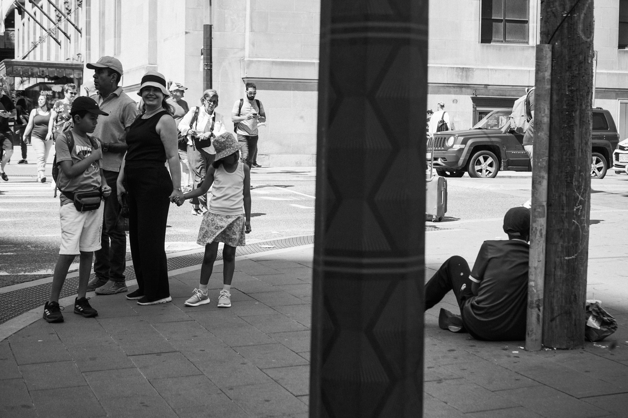 Family waiting on a sidewalk beside a person sitting on the ground, divided by a pillar