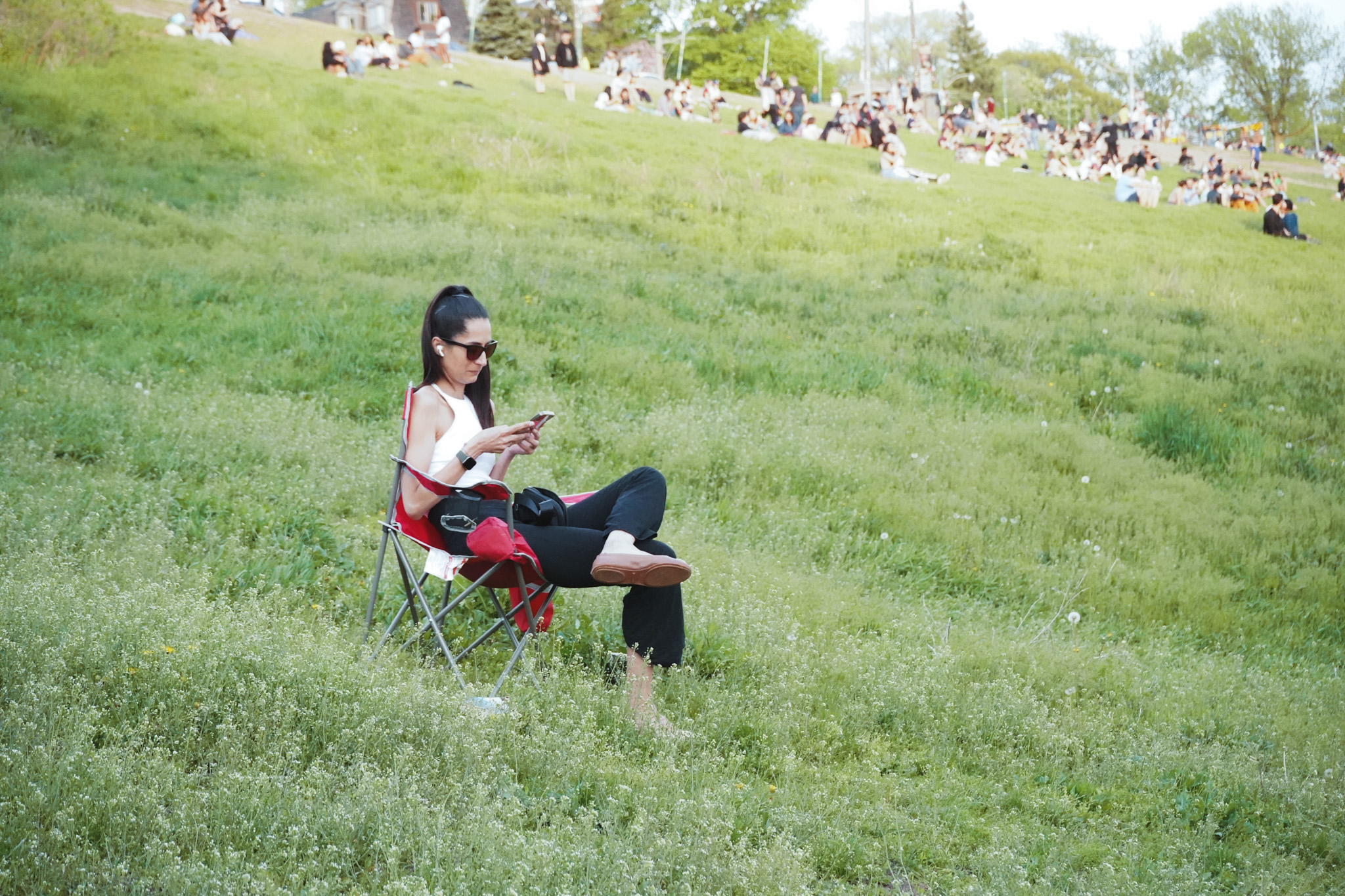 Woman sitting alone in a lawn chair on a grassy hill, phone in hand, crowds behind her