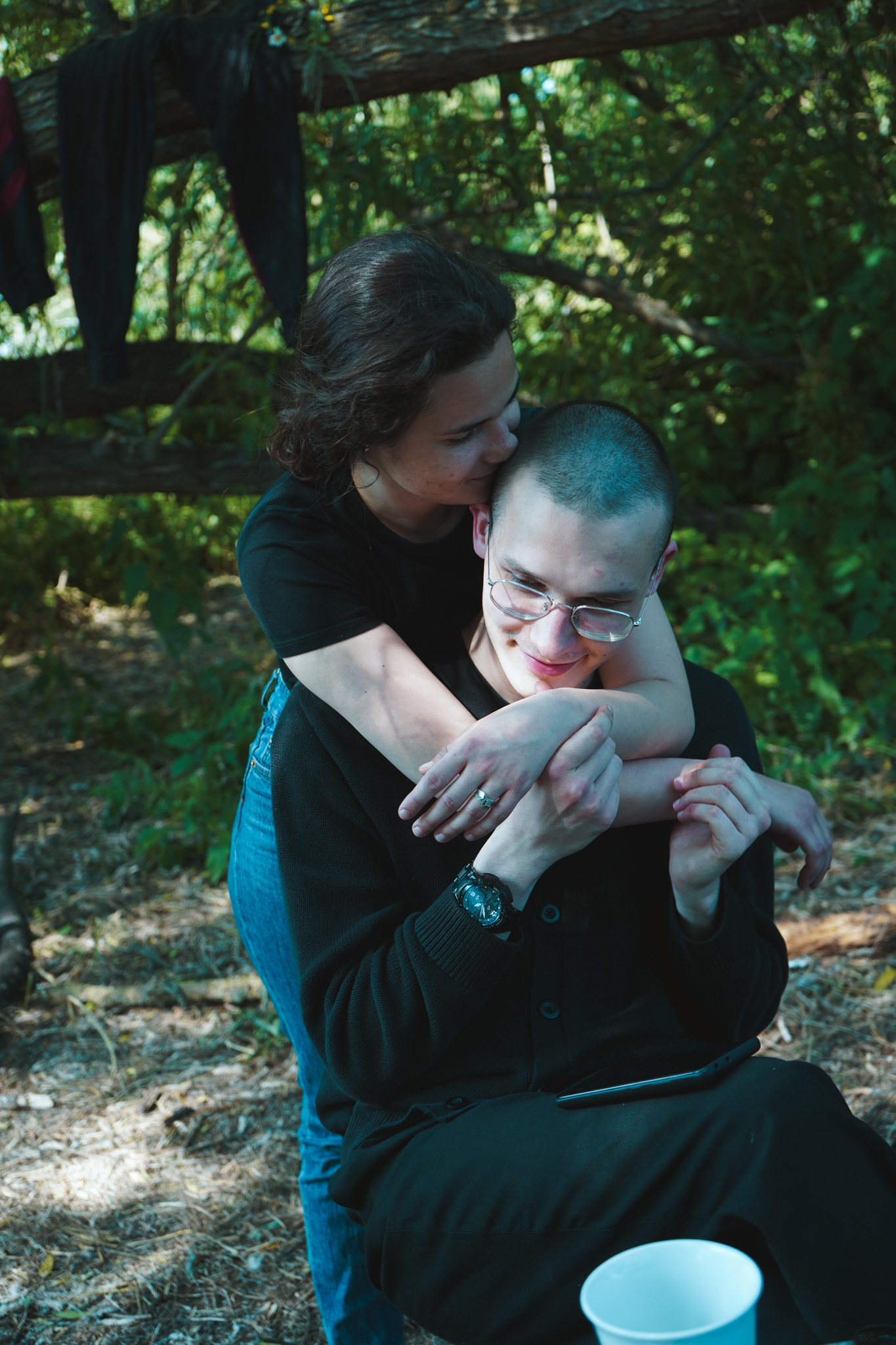 Woman kissing a man on the cheek in a wooded area, arms wrapped around him