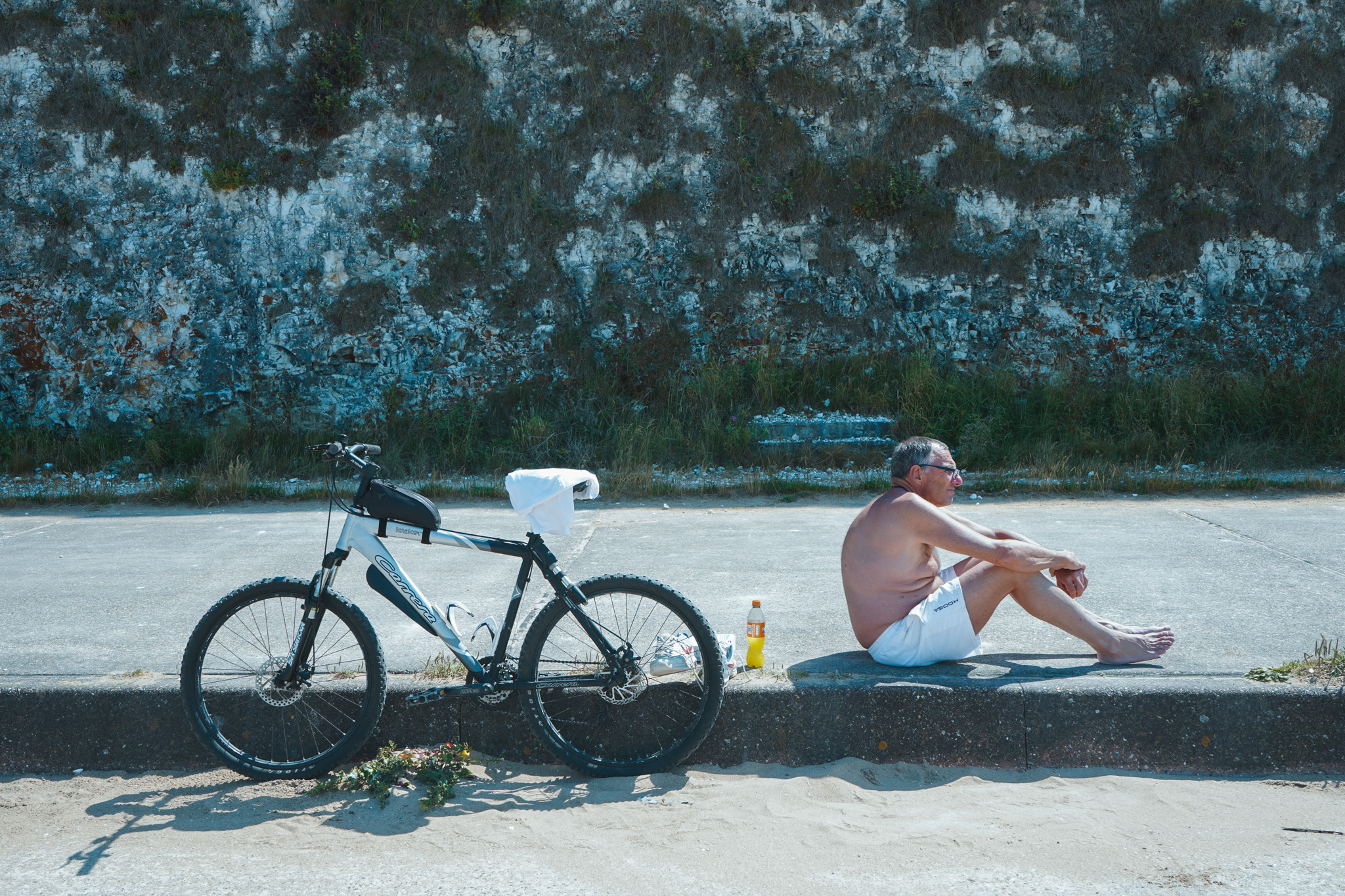 Shirtless man sitting by a curb with his bike, resting against a moss-covered wall