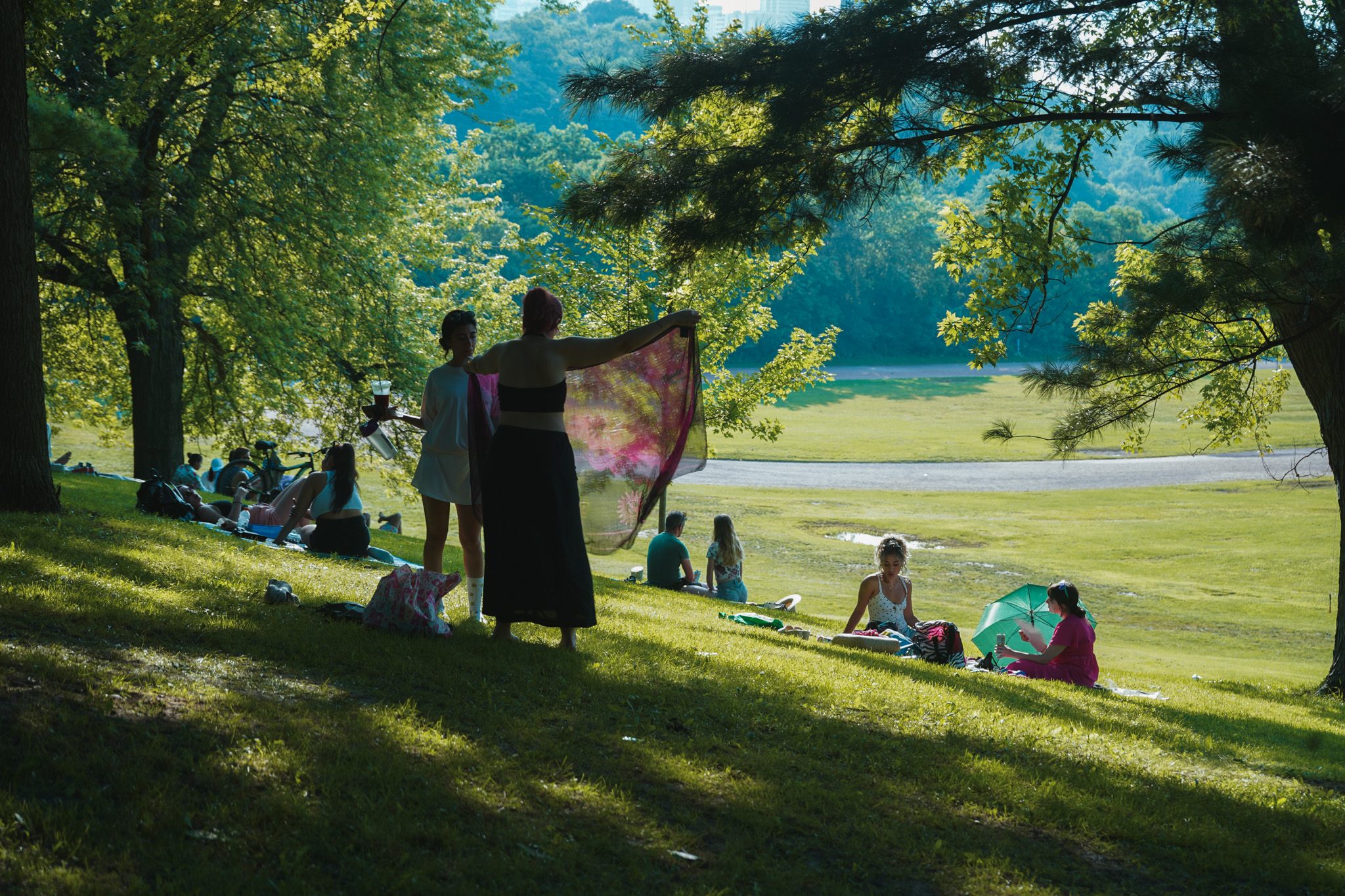 Woman spreading a blanket on a shaded hillside, people lounging on the grass around her
