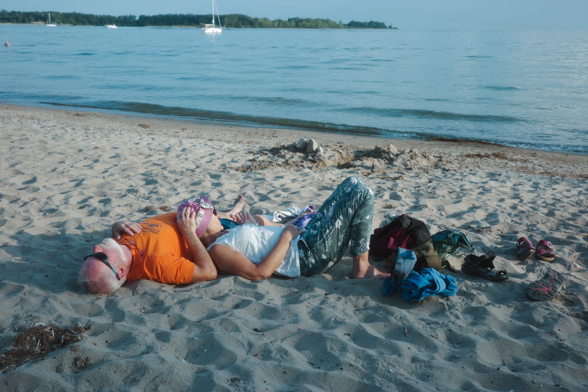 Two people resting on a beach, shoes scattered, water and a sailboat behind